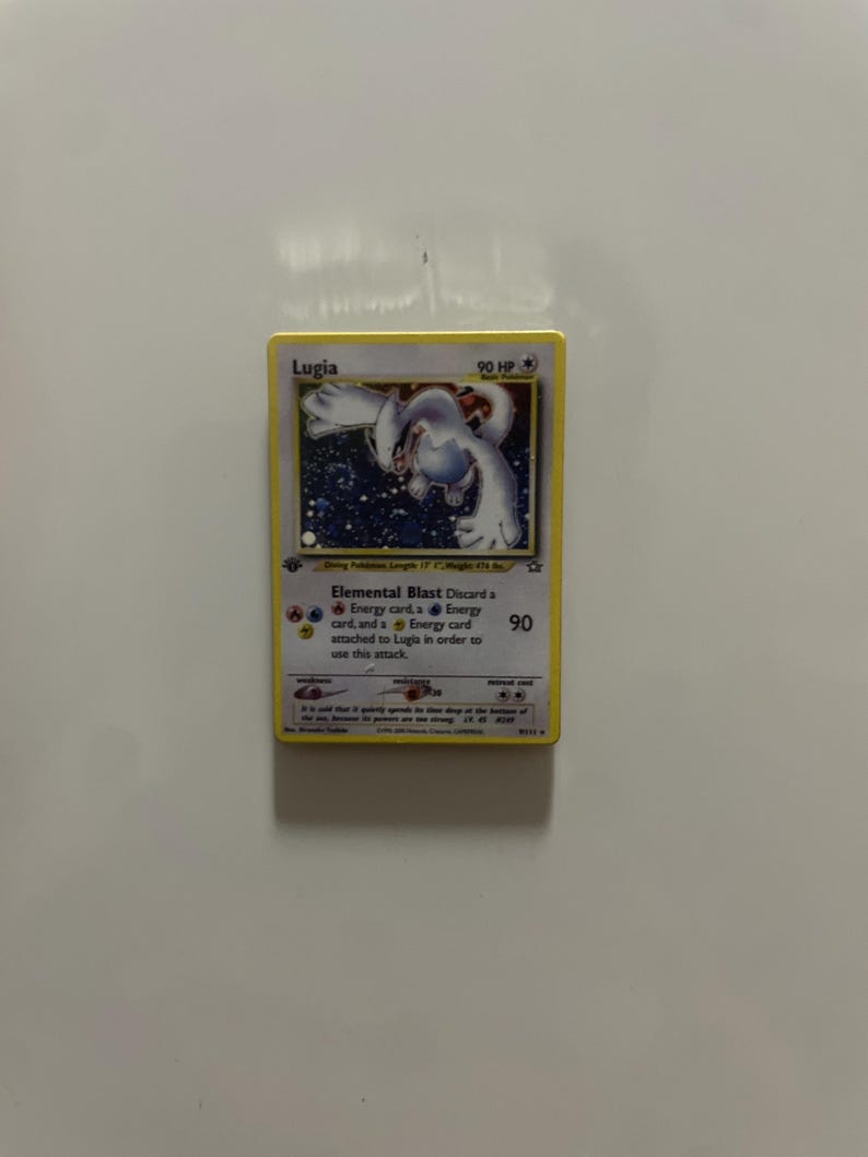 May include: A Lugia trading card featuring a white creature illustration, 90 HP, and the Elemental Blast attack text. The card displays fire, water, and lightning energy symbols, with a yellow border and detailed game statistics printed below.