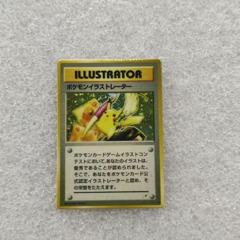May include: A rectangular trading card with a gold border featuring the word ILLUSTRATOR at the top. The central illustration depicts a yellow Pikachu character holding a paintbrush. Japanese text is printed below the image on a gray background.