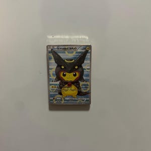May include: A rectangular trading card featuring a yellow Pikachu character wearing a black Umbreon-themed hood. The card has a light blue background with white circular patterns, Japanese text at the top, and game statistics printed on the right side.