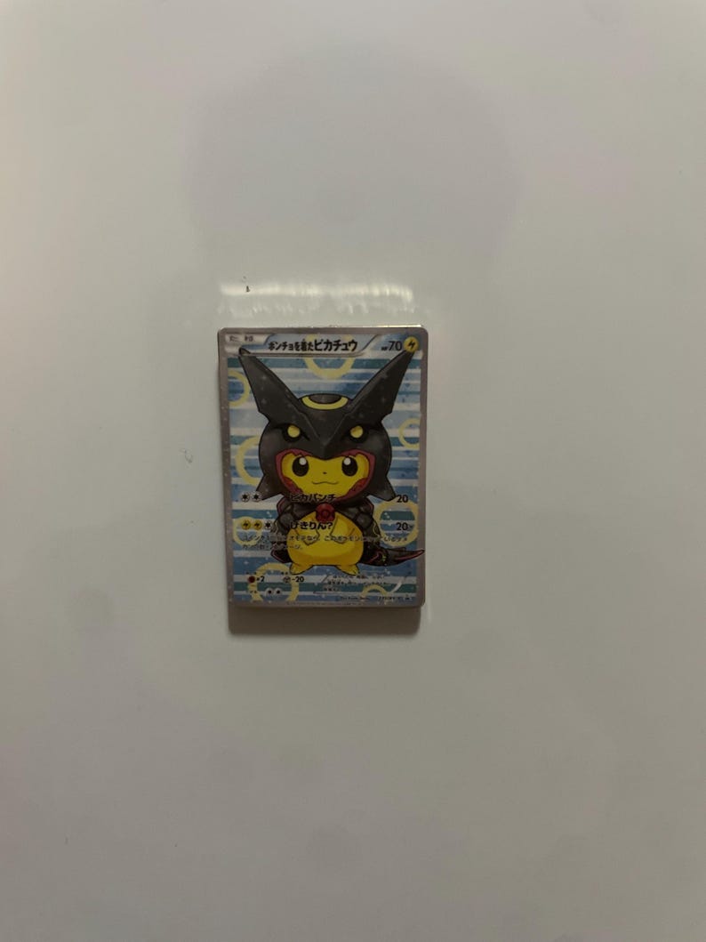 May include: A rectangular trading card featuring a yellow Pikachu character wearing a black Umbreon-themed hood. The card has a light blue background with white circular patterns, Japanese text at the top, and game statistics printed on the right side.
