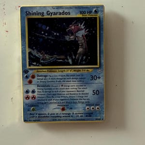May include: A Shining Gyarados trading card with 100 HP, featuring a red sea serpent illustration. The card text includes Outrage and Devastate attacks, with a listed length of 21' 4" and weight of 518 lbs. The border is gold-colored.