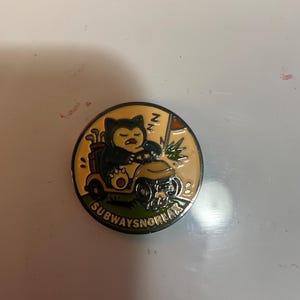 May include: A round enamel pin featuring a dark blue and cream-colored creature driving a yellow golf cart on a green field. The design includes a golf bag, a flag, and the text SUBWAYSNORLAX printed in black along the bottom edge of the pin.
