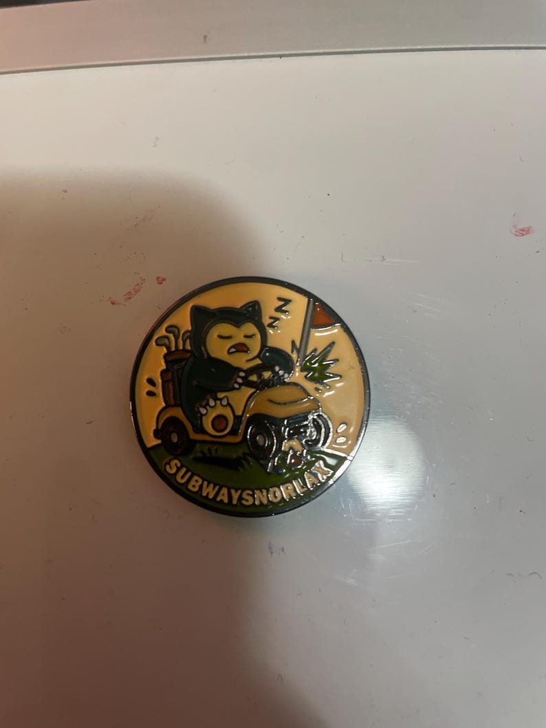 May include: A round enamel pin featuring a dark blue and cream-colored creature driving a yellow golf cart on a green field. The design includes a golf bag, a flag, and the text SUBWAYSNORLAX printed in black along the bottom edge of the pin.