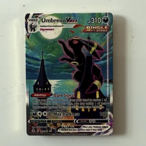 May include: A rectangular Umbreon VMAX trading card featuring a large, dark, rabbit-like creature standing atop a tall, pointed tower under a full moon. The card displays the text Umbreon VMAX, Single Strike, Dark Signal, and Max Darkness 160.