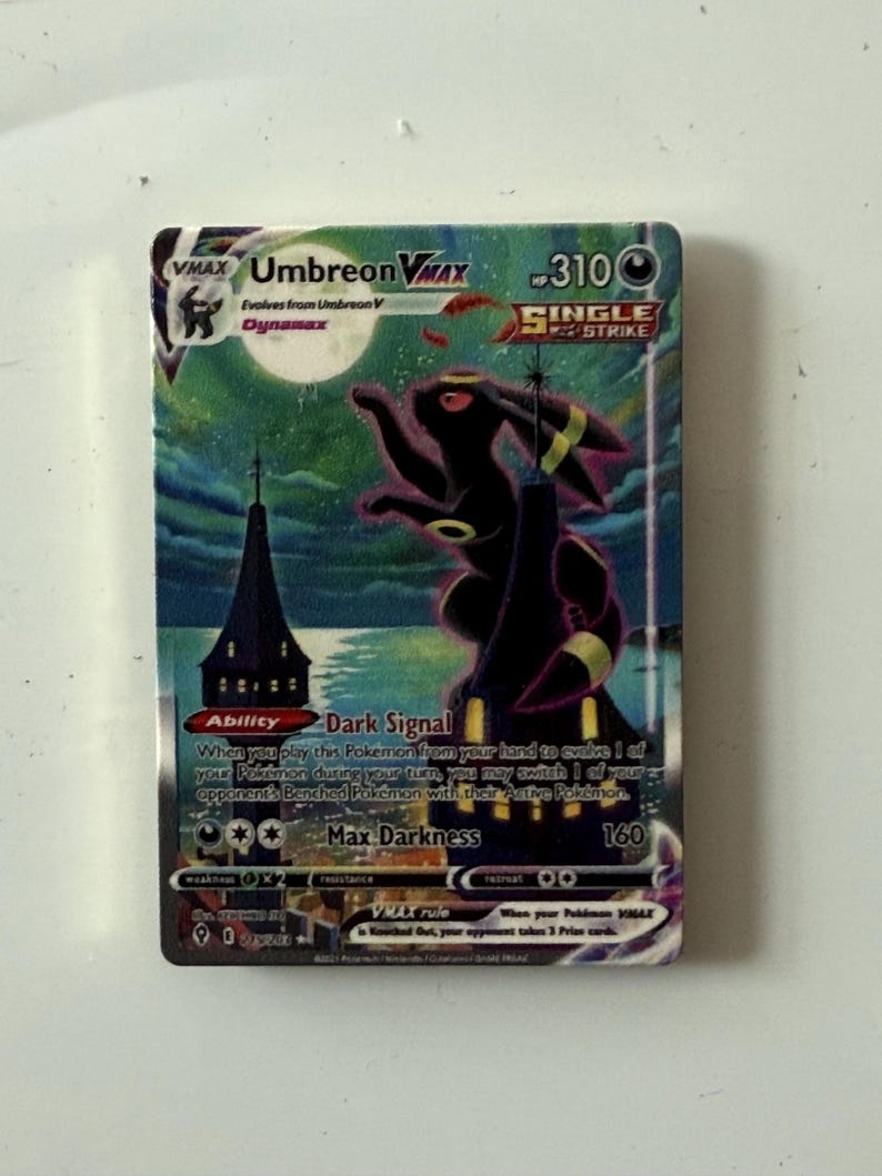May include: A rectangular Umbreon VMAX trading card featuring a large, dark, rabbit-like creature standing atop a tall, pointed tower under a full moon. The card displays the text Umbreon VMAX, Single Strike, Dark Signal, and Max Darkness 160.