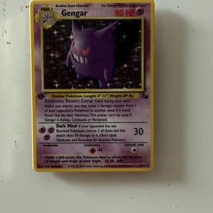 May include: A Gengar trading card featuring a purple ghost-like character with red eyes and a wide grin. The card displays 80 HP, the Pok&eacute;mon Power Curse, and the Dark Mind attack. It includes text detailing the character's length of 4' 11" and weight of 89 lbs.