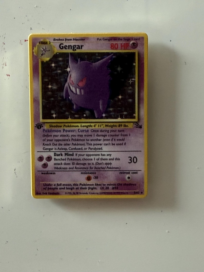 May include: A Gengar trading card featuring a purple ghost-like character with red eyes and a wide grin. The card displays 80 HP, the Pok&eacute;mon Power Curse, and the Dark Mind attack. It includes text detailing the character's length of 4' 11" and weight of 89 lbs.