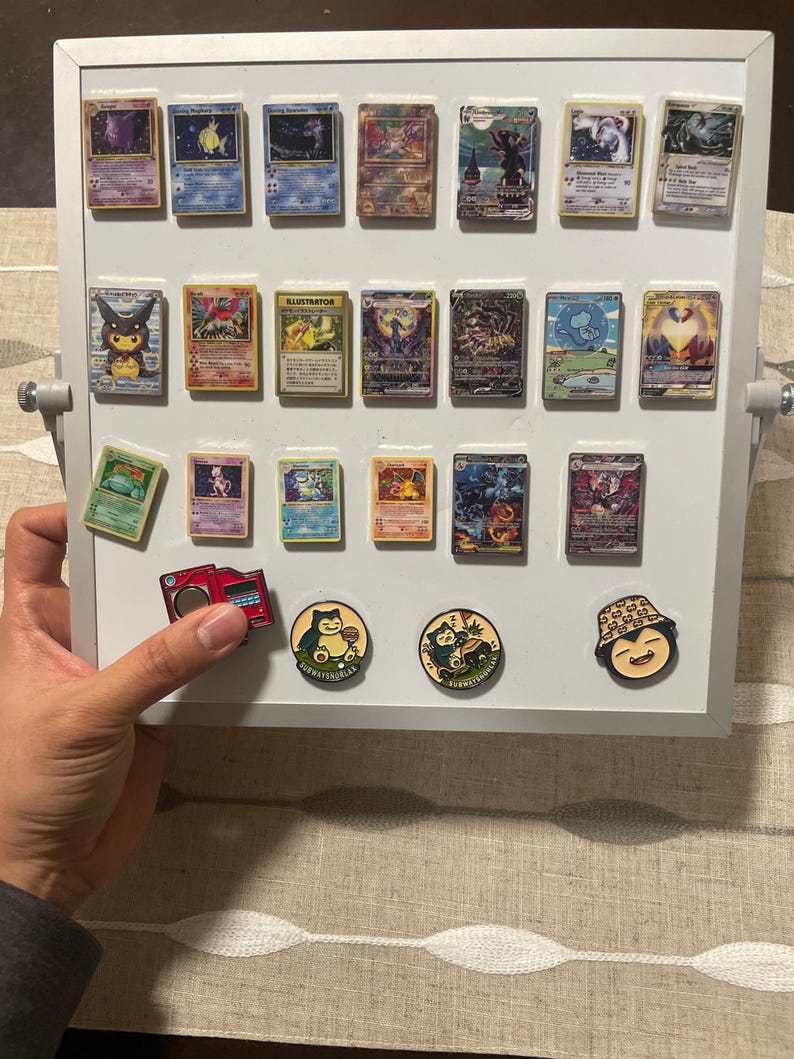 May include: A white magnetic display board featuring seventeen rectangular trading cards arranged in three rows, with three circular enamel pins positioned below the cards. The pins depict Snorlax characters and a red electronic device.