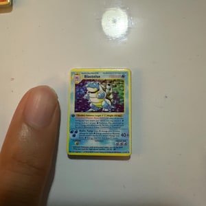 May include: A rectangular Blastoise trading card featuring a blue turtle-like creature with cannons on its back. The card has a yellow border, blue background, and text detailing the character's name, HP, and special moves like Hydro Pump.