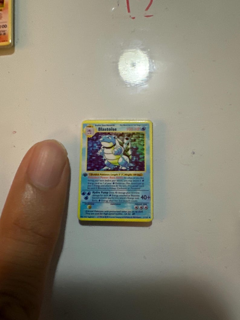 May include: A rectangular Blastoise trading card featuring a blue turtle-like creature with cannons on its back. The card has a yellow border, blue background, and text detailing the character's name, HP, and special moves like Hydro Pump.