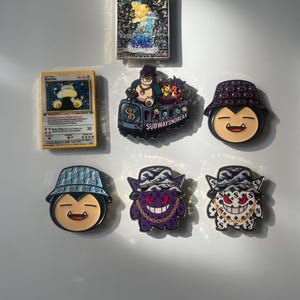 May include: A collection of items including a Snorlax trading card, a holographic card, and five enamel pins. The pins feature cartoon characters wearing bucket hats, including a subway train design with the text SUBWAYSNORLAX and various patterns.