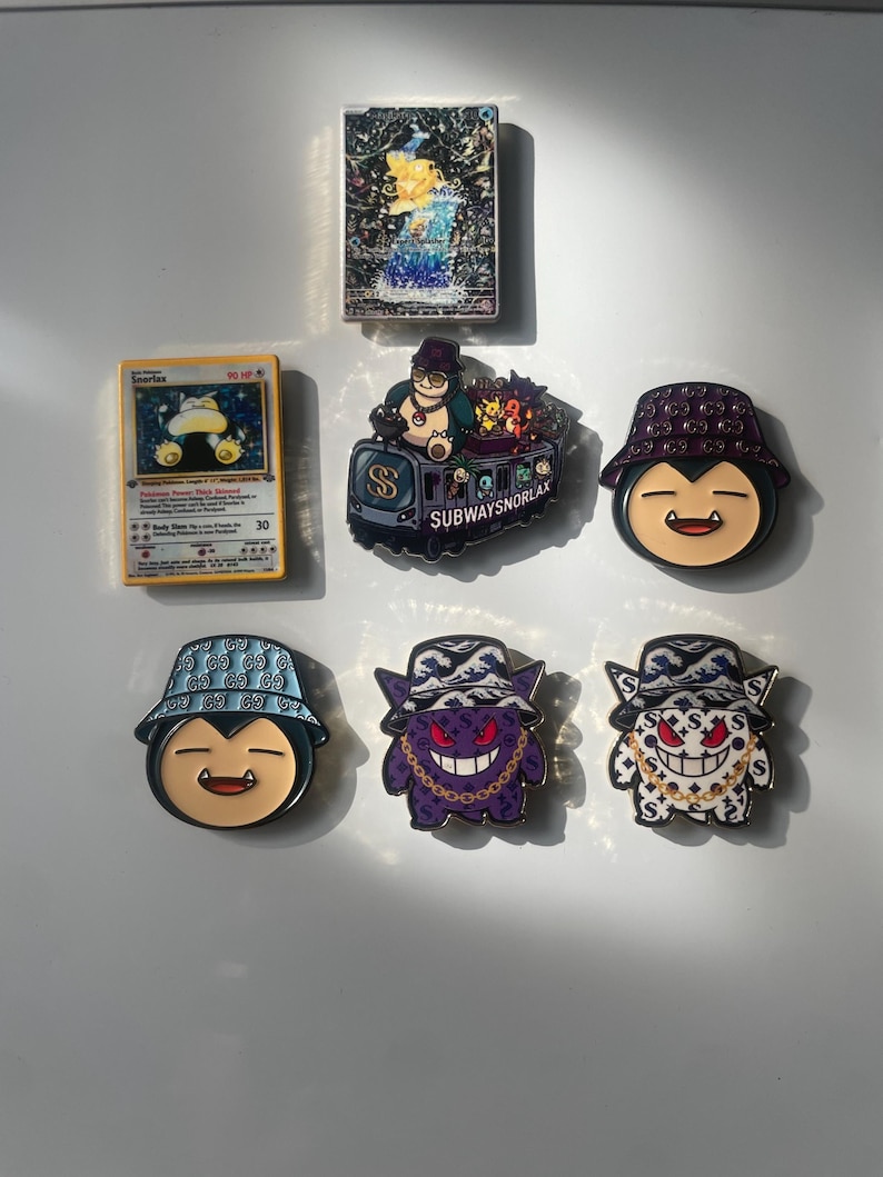 May include: A collection of items including a Snorlax trading card, a holographic card, and five enamel pins. The pins feature cartoon characters wearing bucket hats, including a subway train design with the text SUBWAYSNORLAX and various patterns.