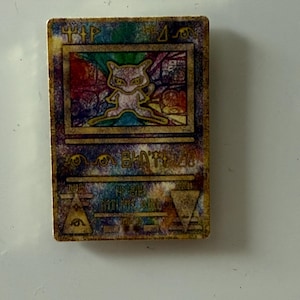 May include: A rectangular trading card featuring a central illustration of a small, light-colored creature with large eyes. The card has a weathered, multi-colored background with gold-toned borders and various abstract symbols and text printed on it.