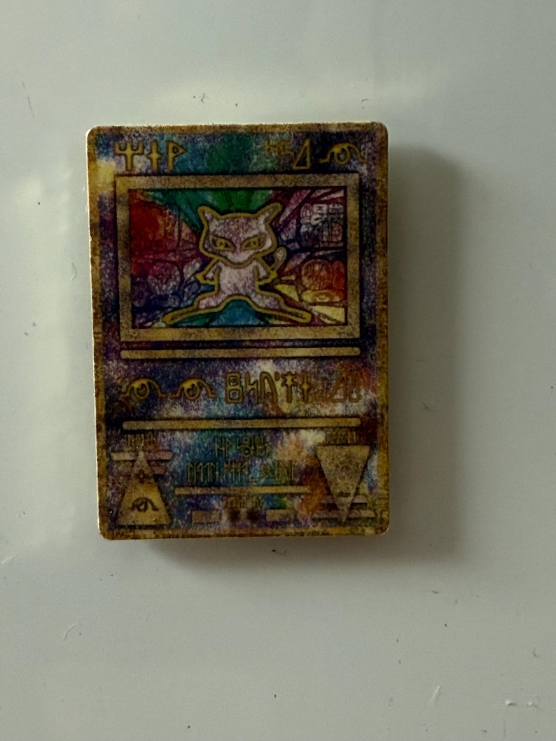 May include: A rectangular trading card featuring a central illustration of a small, light-colored creature with large eyes. The card has a weathered, multi-colored background with gold-toned borders and various abstract symbols and text printed on it.