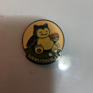 May include: A circular enamel pin featuring a cartoon creature with a cream-colored belly and dark blue fur, holding a brown hamburger. The background is a pale yellow circle above a green patch of grass. The text SUBWAYSNORLAX is printed in black.