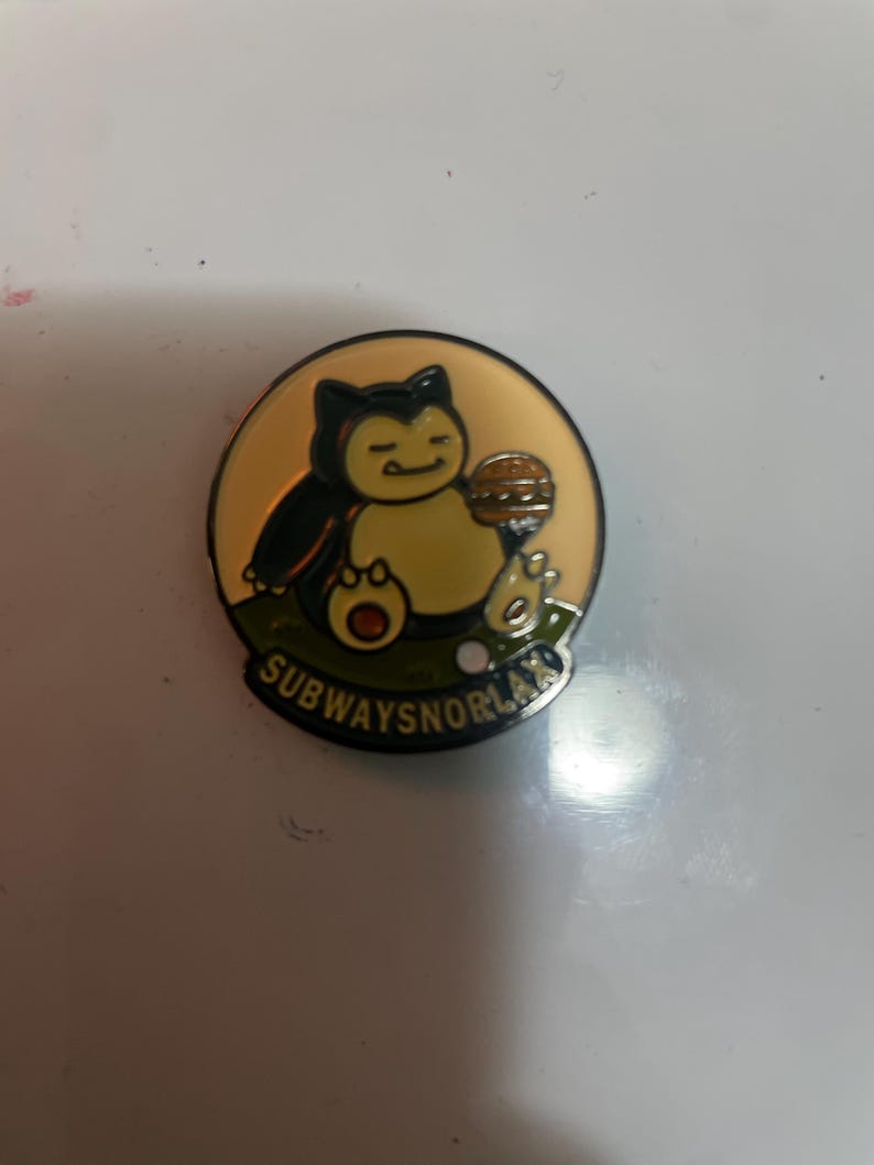 May include: A circular enamel pin featuring a cartoon creature with a cream-colored belly and dark blue fur, holding a brown hamburger. The background is a pale yellow circle above a green patch of grass. The text SUBWAYSNORLAX is printed in black.
