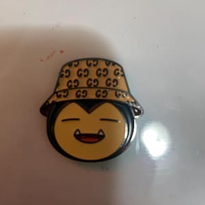 May include: A metal enamel pin featuring a cartoon character face with closed eyes and visible fangs, wearing a tan bucket hat patterned with a repeating black logo design. The character has a light yellow face and a dark outline.