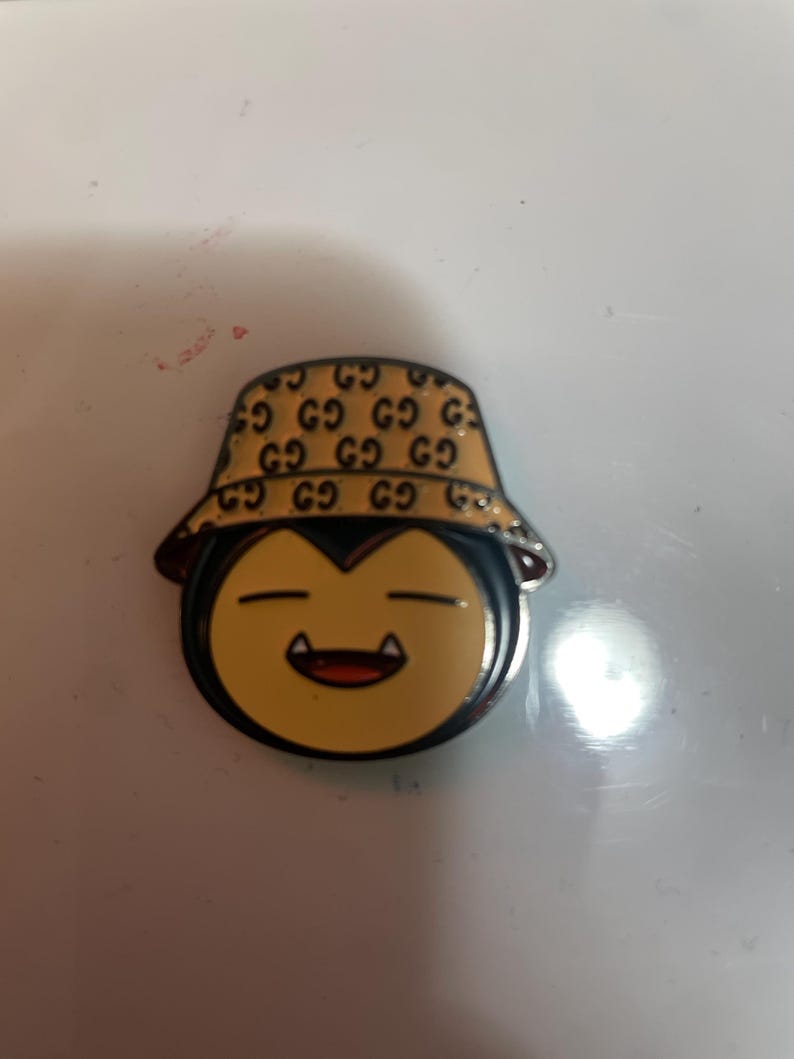 May include: A metal enamel pin featuring a cartoon character face with closed eyes and visible fangs, wearing a tan bucket hat patterned with a repeating black logo design. The character has a light yellow face and a dark outline.