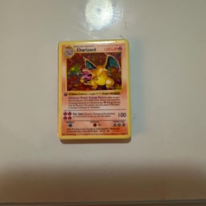May include: A holographic Charizard trading card featuring the orange dragon-like creature with wings spread, displaying 120 HP, the Energy Burn Pok&eacute;mon Power, and the Fire Spin attack with a damage value of 100, printed with yellow borders.