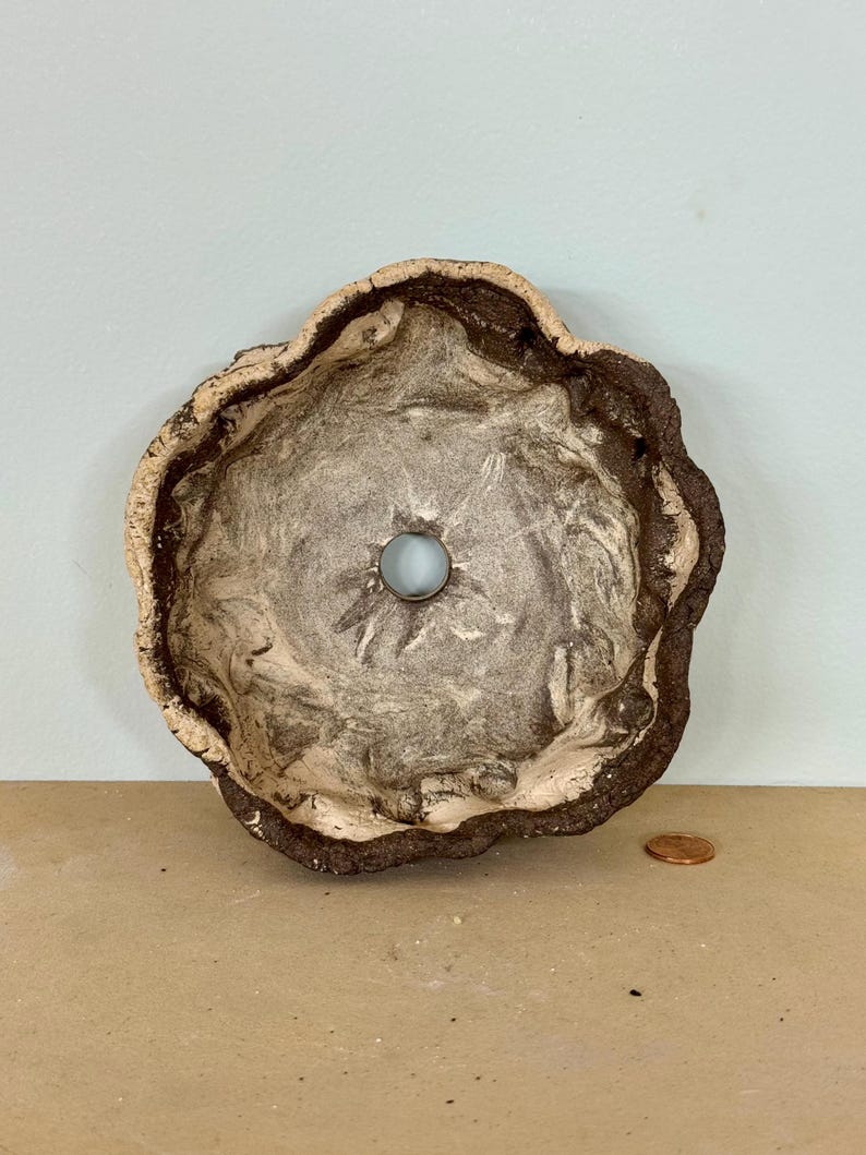 May include: A handmade ceramic bowl with a unique, organic shape. The bowl features a light brown rim, a dark brown exterior, and a marbled interior with a light blue glass bead in the center. The bowl is approximately 6 inches in diameter.