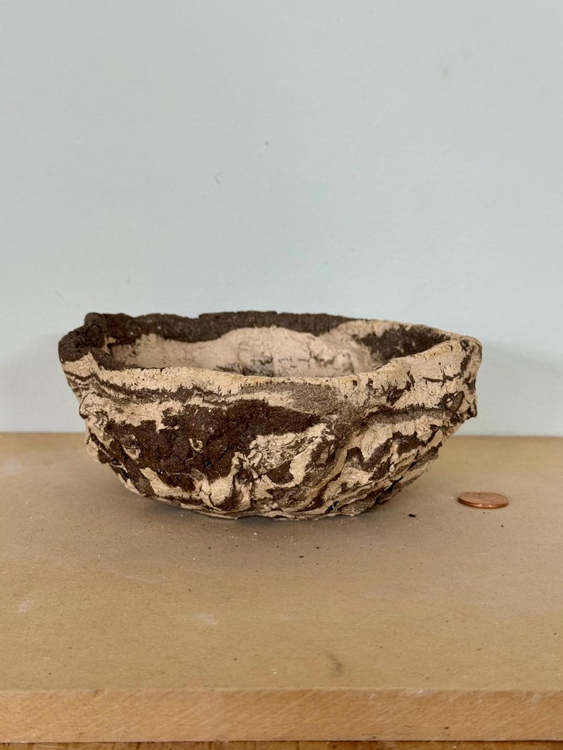 May include: A small, handmade bowl with a textured, organic appearance. The bowl features a dark brown and beige marbled pattern. The rim has a rough, uneven edge. A penny is next to the bowl for size reference.