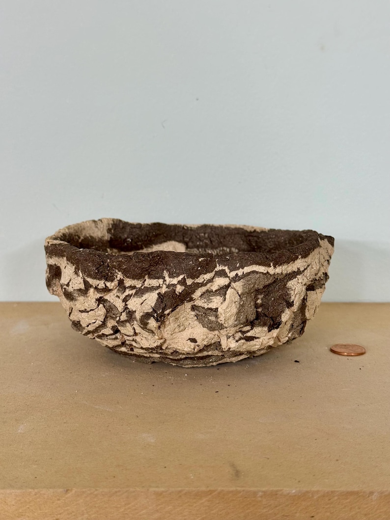 May include: A small, handmade bowl with a textured, organic appearance. The bowl features a light beige base with dark brown accents, creating a rustic aesthetic. A penny is next to the bowl for size reference.