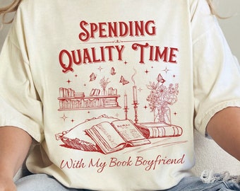 May include: A white t-shirt with a red graphic design that says "Spending Quality Time With My Book Boyfriend". The graphic features a bookshelf with books, candles, and flowers.