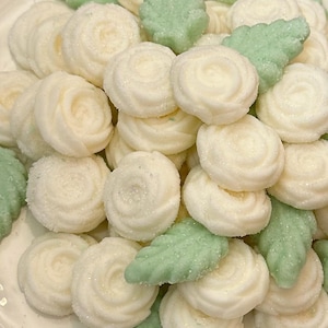 May include: A plate of white, rose-shaped candies and green leaf-shaped candies. The candies are arranged in a pile on a white plate with a floral border. The candies appear to be sugared.