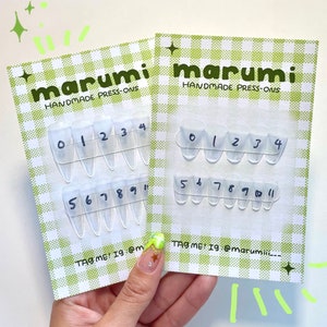 Op de afbeelding: Twee kaarten met doorzichtige, genummerde persnagels. Elke kaart heeft de merknaam "marumi" en "HANDMADE PRESS-ONS" erop gedrukt. De nagels zijn gerangschikt in rijen, genummerd van 0-4 en 5-11. De achtergrond is een groen-wit geruit patroon.
