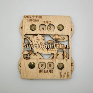 Puede incluir: Un token de Saproling de madera con diseños de hongos y árboles cortados con láser. El token tiene remaches de color latón y el texto "TOKEN CREATURE SAPROLING" y "SAPROLING TOKEN". El token tiene números para los valores "TAPPED" y "UN-TAPPED".