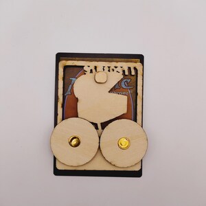 May include: A wooden card holder with two round wooden knobs. The card holder is designed to hold a Magic: The Gathering card. The card holder is made of light brown wood and has a black border.