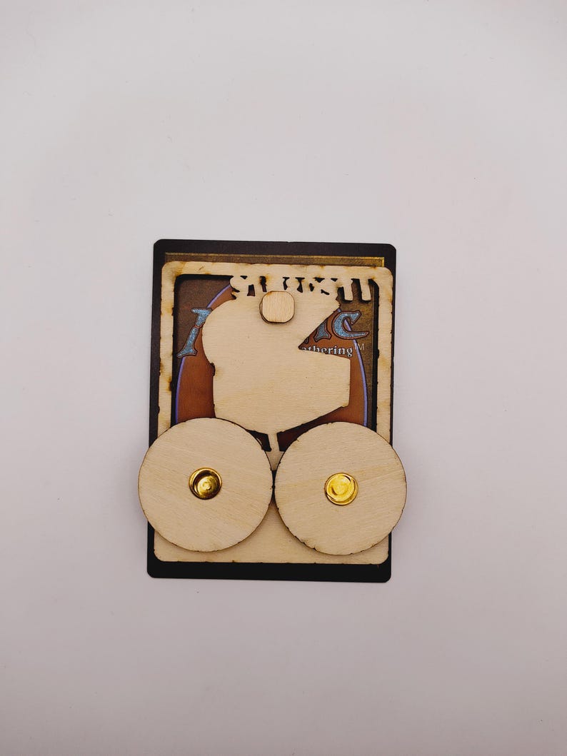 May include: A wooden card holder with two round wooden knobs. The card holder is designed to hold a Magic: The Gathering card. The card holder is made of light brown wood and has a black border.