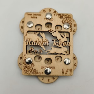 May include: A wooden Rabbit Token for tabletop games. The token features a laser-cut design with the words "Rabbit Token" and rabbit silhouettes. It has a dial for tracking game stats and the text "TAPPED" and "UN-TAPPED".
