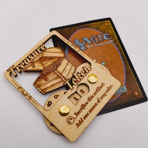May include: A wooden treasure token for Magic: The Gathering. The token features a wooden chest with a sliding lid and a counter that can be set to 1, 5, or 8. The text on the token reads "Treasure Token" and "Sacrifice this artifact: Add one mana of any color."