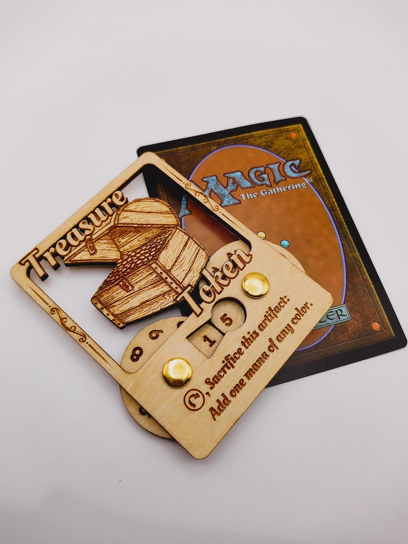 May include: A wooden treasure token for Magic: The Gathering. The token features a wooden chest with a sliding lid and a counter that can be set to 1, 5, or 8. The text on the token reads "Treasure Token" and "Sacrifice this artifact: Add one mana of any color."