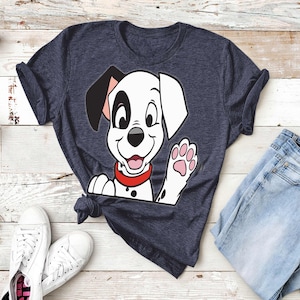 May include: Navy blue t-shirt featuring a cartoon Dalmatian puppy waving with a pink paw. The puppy has black spots, a red collar, and a black ear. The shirt is paired with blue jeans and white sneakers.