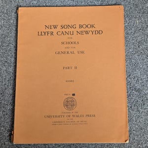 Op de afbeelding: Een vintage songboek met een bruine kaft. De titel luidt "NEW SONG BOOK LLYFR CANU NEWYDD" met extra tekst voor scholen en algemeen gebruik. De kaft bevat ook "DEEL II" en "SCORE". Uitgegeven door de University of Wales Press.