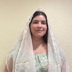 May include: A white sheer scarf with a floral pattern in a light brown colour. The scarf is draped over a person's head and shoulders.