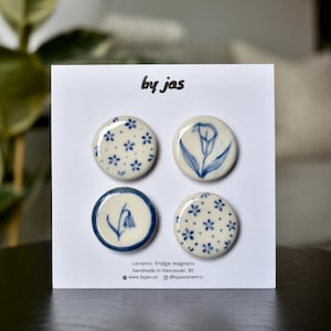 Puede incluir: Un juego de cuatro imanes de cerámica redondos para nevera sobre una tarjeta de presentación blanca con la etiqueta by jas. Los imanes presentan diseños pintados a mano en azul y blanco, incluyendo dos con motivos florales, un lirio y una campanilla.