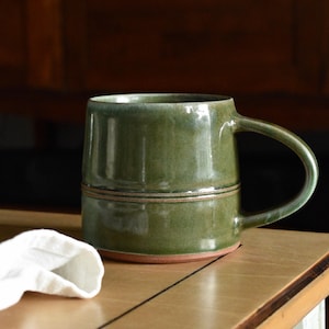 May include: A cylindrical ceramic mug with a glossy olive green glaze and a smooth finish. The mug features a prominent horizontal ridge around the centre and a curved handle on the right side. The base reveals a natural, unglazed terracotta clay rim.
