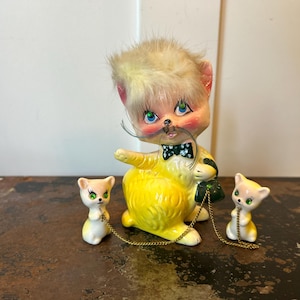 May include: A ceramic cat figurine with yellow fur, green rhinestone eyes, and a black bow tie. The cat has a tuft of blonde faux fur on its head, long black whiskers, and is connected by gold chains to two smaller matching cat figurines.