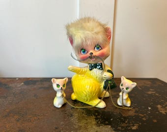 Puede incluir: Una figura de gato de cerámica con pelaje amarillo, ojos de estrás verdes y una pajarita negra. El gato tiene un mechón de pelo sintético rubio en la cabeza, largos bigotes negros y está unido por cadenas doradas a dos figuras de gatos más pequeñas.