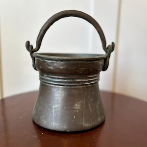 May include: A small, antique copper bucket with a curved handle. The bucket has a rounded base and a slightly flared body. The handle is attached to the bucket with decorative brackets. The metal has a dark, aged patina, suggesting its vintage. This decorative piece is ideal for home decor.