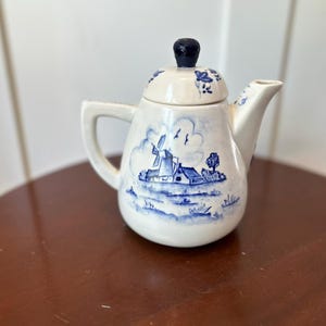May include: A small, white ceramic teapot with a blue windmill and landscape design. The lid has a black knob and blue floral accents. The teapot has a handle and a spout, and is likely for serving tea.