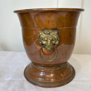 May include: A polished copper planter with a wide, flared rim and a circular base. The exterior features a prominent brass lion head ornament centered on the front, which holds a circular brass ring handle. The metal shows signs of natural patina.