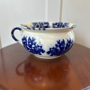 May include: A white and blue ceramic chamber pot with a handle. The pot features a blue floral pattern around the body and rim. The chamber pot is sitting on a brown wooden table.