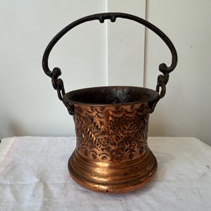 May include: A copper bucket featuring a dark metal swing handle and an embossed pattern along the exterior. The vessel has a wide, flared base and a cylindrical body with a textured, repeating design of arches and leaf-like motifs in metallic copper.