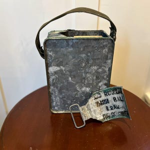 May include: A weathered, rectangular metal container with a handle, possibly a vintage ammunition box. The container is gray with signs of wear and tear. A detached metal piece with text lies nearby.