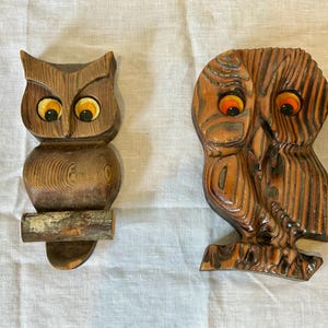 May include: Two carved wooden owl figurines. The left owl features a smooth, light brown wood grain with large yellow eyes and sits on a small branch. The right owl has a darker, textured wood grain with prominent orange eyes and a carved wing.