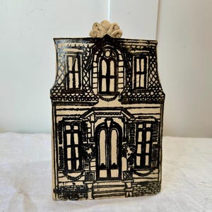 May include: A cream-colored ceramic house figurine featuring detailed black line drawings of a two-story building with a central arched doorway, multiple windows, and a decorative roof ornament. The structure has a flat front and rectangular shape.